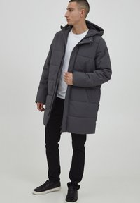 Grå polstret kåpe med hette, med trykknapper og glidelås, sidelommer og quiltet design. Brukt sammen med en hvit skjorte og svarte bukser.