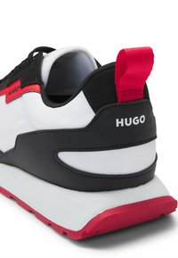 Black and white sneaker with red accents. Features a textured fabric upper, padded collar, and a rubber outsole. "HUGO" branding on heel.
