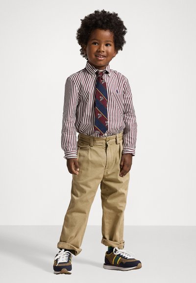 Svītraina sarkana un balta krekla jaka, tumši zila kaklasaite ar rakstainiem akcentiem, haki krāsas bikses un daudzkrāsainas kedas ar baltām auklām.