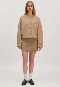 Geknipte beige jas met grote knopen, enkel borstzakje; gecombineerd met een aansluitende beige gebreide rok. Zwarte loafers en witte sokken maken de look compleet.