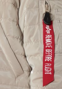 Kurtka pikowana z połyskującej beżowej tkaniny z pikowanym wzorem. Posiada czerwoną metkę z białym napisem "REMOVE BEFORE FLIGHT" oraz srebrne zamki błyskawiczne.