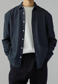 Chemise bleu marine foncé à boutons avec un tissu texturé, une poche poitrine unique et des boutons blancs, portée par-dessus un t-shirt gris clair et un pantalon noir.