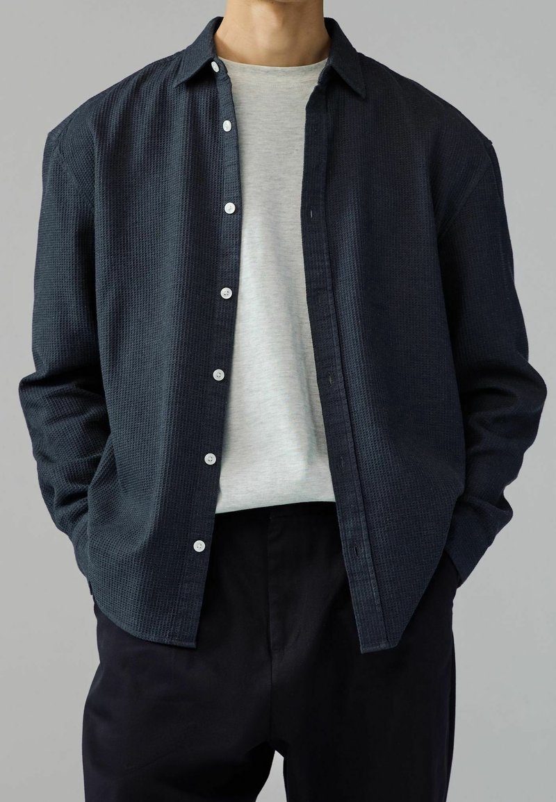 Chemise bleu marine foncé à boutons avec un tissu texturé, une poche poitrine unique et des boutons blancs, portée par-dessus un t-shirt gris clair et un pantalon noir.