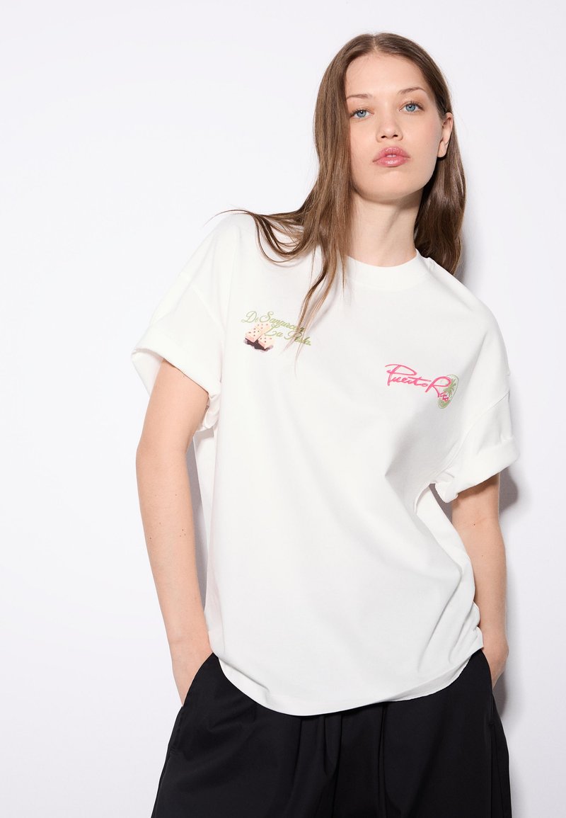 Jeune femme aux longs cheveux bruns portant un t-shirt blanc à motif graphique et un pantalon noir, debout les mains dans les poches sur fond blanc.