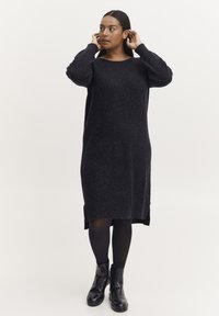 Rochie tricotată supradimensionată, de culoare închisă, cu mâneci lungi, decolteu rotund, slituri laterale și o textură moale, asortată cu botine negre până la gleznă.