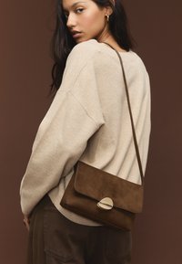 Smešnjava rjava torbica iz usnja z zaponko na preklop in zlato detajlom. Model nosi bež pulover, s trakcem čez ramo in teksturirano površino.