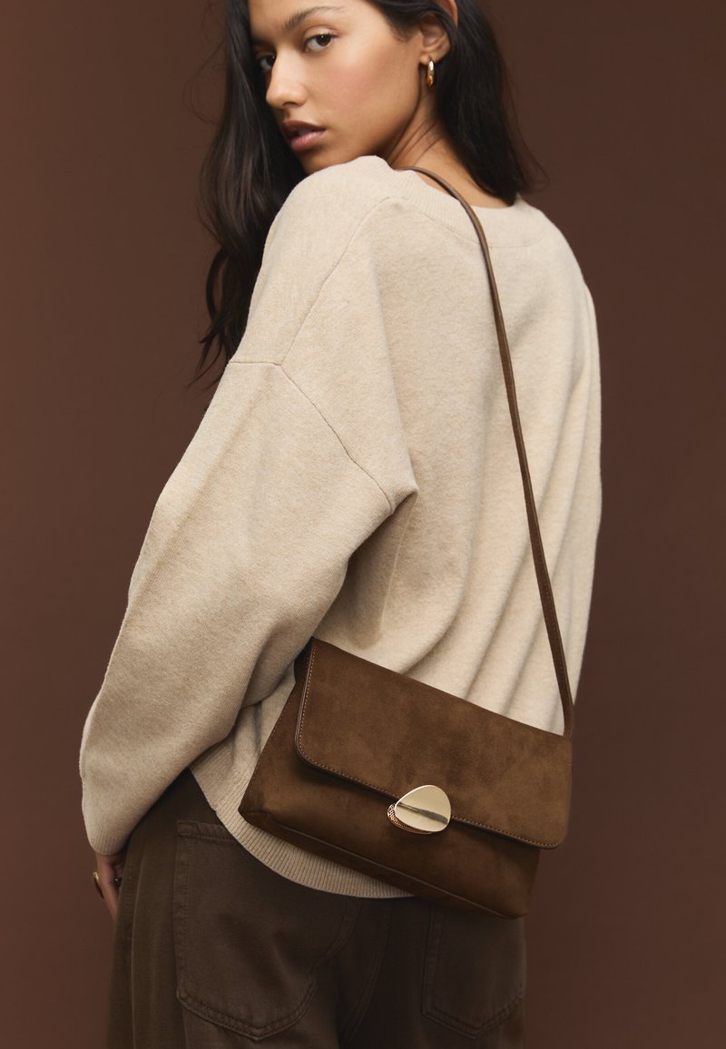 Smešnjava rjava torbica iz usnja z zaponko na preklop in zlato detajlom. Model nosi bež pulover, s trakcem čez ramo in teksturirano površino.
