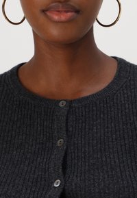 Cardigan côtelé gris foncé avec un col rond et cinq fermetures par boutons, complété par des boucles d'oreilles rondes en or.