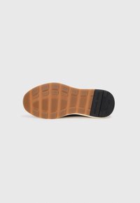 Shoe sole with a textured gum rubber bottom, featuring a grid pattern and logo detail. Colors include tan, black, and off-white.