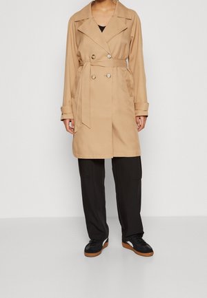 Person trägt einen knie-langen beigen Trenchcoat mit gebundenem Gürtel, schwarze Hose und schwarze Sneakers mit weißen und braunen Sohlen, steht an einer weißen Wand.