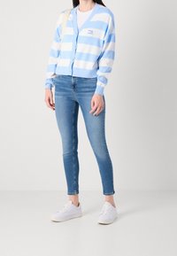 Cárdigan a rayas azul y blanco con botones, llevado sobre una camiseta blanca y combinado con jeans ajustados azules y zapatillas blancas.