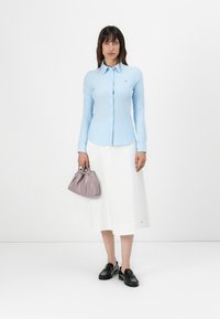 Camicia azzurra a righe con bottoni, gonna bianca e mocassini neri. Tiene in mano una piccola borsa lavanda. Tessuto morbido, design aderente.