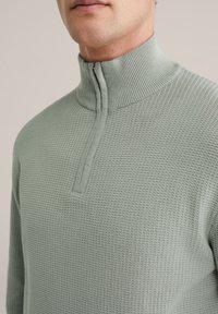 Svijetlozeleni pulover od teksturiranog pletenog materijala s visokim ovratnikom i poluzatvaračem. Ima geometrijski uzorak za dodatni detalj.