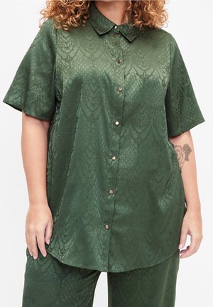 Vrouw draagt een groen satijnen kortemouwenoverhemd met knopen en bijpassende broek met subtiel slangenhuidpatroon, tatoeage zichtbaar op de onderarm.