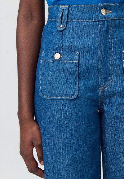 Jean en denim bleu taille haute avec une poche avant et un bouton doré, des coutures contrastées et une texture lisse.