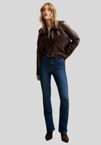 Giacca corta in pelle marrone con cuciture bianche, blusa scura, jeans blu e stivali alla caviglia scuri, presentata su uno sfondo grigio.