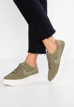Personne portant des baskets en daim vert olive avec des semelles blanches épaisses et un jean noir à ourlet effiloché sur un fond blanc.