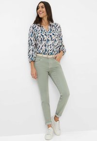 Gemusterte Bluse in Blau- und Rosatönen mit V-Ausschnitt. Hellgrüne, eng zulaufende Hose mit umgeschlagenen Säumen, kombiniert mit weißen Turnschuhen.