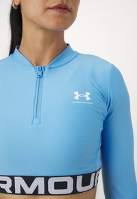 Heledsinine poolrihmaga pika varrukaga topp, mis on valmistatud venivast materjalist. Rinnal on valge Under Armouri logo ja must vöökoht, millel on tekst.