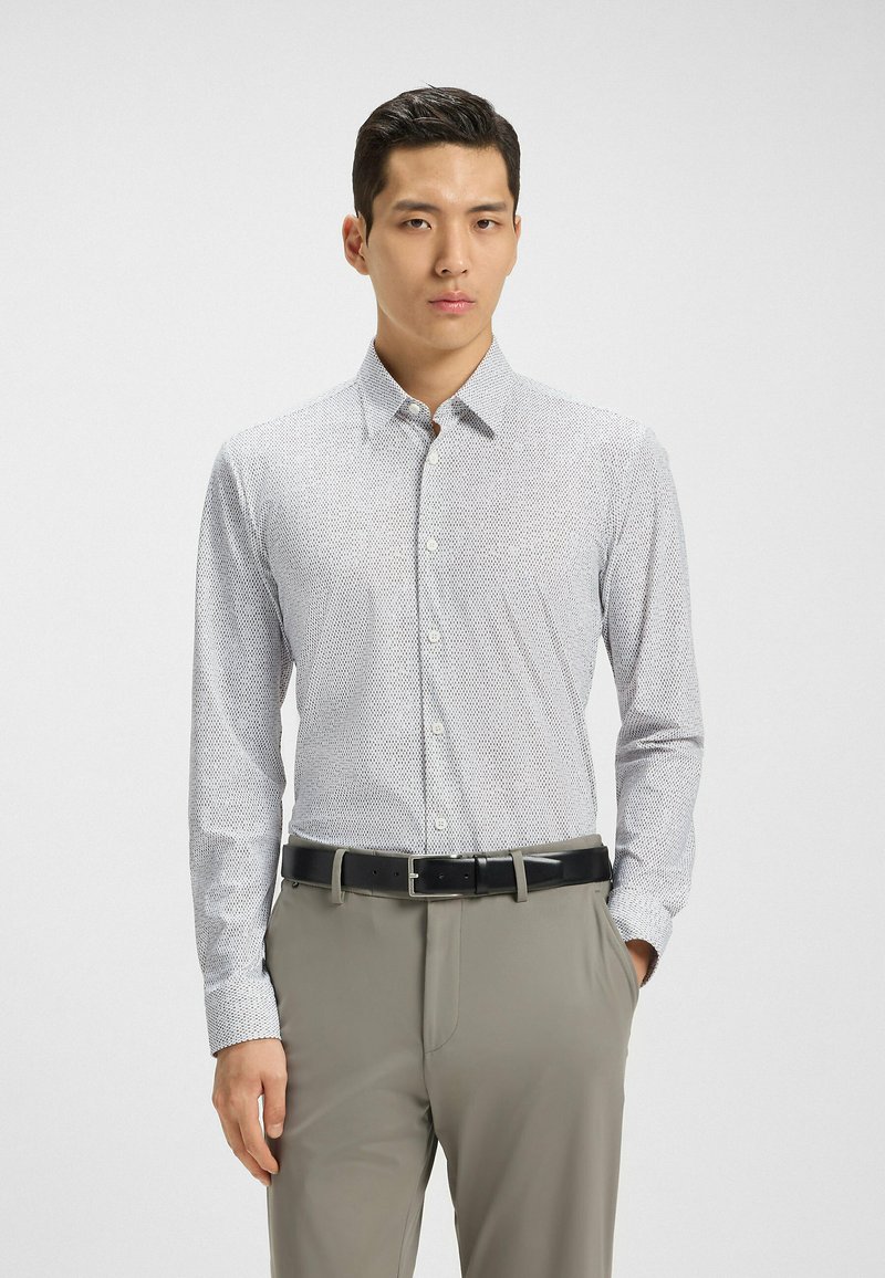 Biała koszula z długim rękawem, zapinana na guziki, z delikatnym niebieskim wzorem, dopasowany krój i kołnierz typu spread; zestawiona z jasnobeżowymi spodniami i czarnym paskiem.