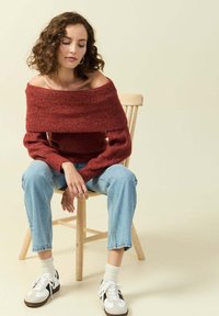 Maglione rosso scollato sulle spalle con texture a coste, abbinato a jeans blu chiaro a gamba dritta e scarpe bianche con accenti neri, seduto su uno sgabello di legno.