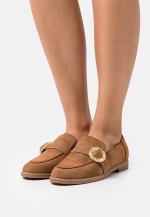 Mocassins en cuir suédé marron avec des boucles en écaille de tortue rondes, portés pieds nus, présentés sur un fond blanc uni.