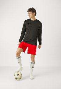 Langärmliges schwarzes Trikot, rote Shorts, weiße Socken mit schwarzen Streifen und weiße Fußballschuhe, steht mit einem Fuß auf einem mehrfarbigen Fußball.