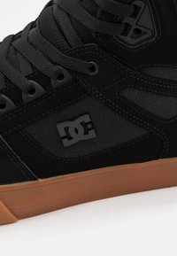 Black high-top sneaker made of synthetic material with a rubber sole, featuring a textured fabric panel and a prominent logo on the side.