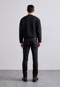 Black sweatshirt with round neck, paired with black fitted jeans. Model stands with back to the camera, wearing brown shoes. Simple design.