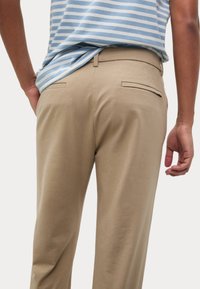 I pantaloni beige presentano una texture liscia, una vestibilità su misura e una tasca posteriore con un'apertura discreta. Abbinati a una camicia a righe blu chiaro.