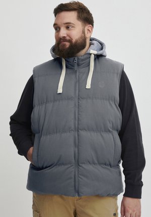 Grå polstret vest med glidelås foran, snorhette og logodetalj. Båret over en svart langermet skjorte, har sidelommer.