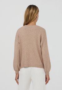 Donna con capelli lunghi che indossa orecchini ad anello esagonali, un maglione beige e pantaloni bianchi, vista di spalle su uno sfondo semplice.