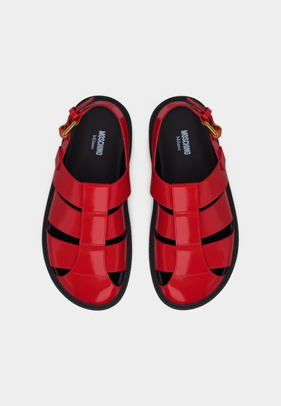Κόκκινα σανδάλια από δερμάτινο λουστρίνι με μαύρες σόλες, ανοιχτή μύτη, πολλαπλές λουρίδες, ρυθμιζόμενο αγκράφα και λογότυπο "Moschino Milano" στο εσωτερικό.