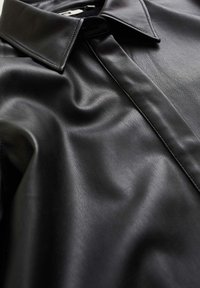 Närbild av en slät svart läderskjorta med spetsig krage och synliga söm detaljer.