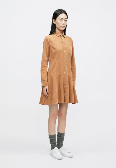 Νεαρή γυναίκα που φοράει καφέ φόρεμα με μακριά μανίκια και κουμπιά, γκρι σούρες κάλτσες και λευκά αθλητικά παπούτσια, στέκεται μπροστά σε λευκό φόντο.