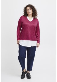 Burgundy långärmad topp lager på lager över en vit skjorta med krage, tillsammans med mörkblå uppvikta jeans och svarta skor, med en v-ringad design.