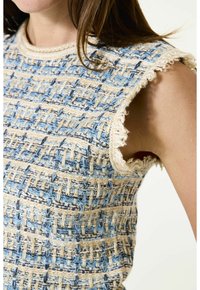 Close-up van een mouwloze gestructureerde gebreide top met een blauw, beige en crèmekleurig geweven patroon en rafelige randen bij de armsgaten en de halslijn.