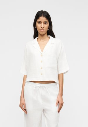 Γυναίκα με μακριά σκούρα μαλλιά φορά χαλαρό, λευκό πουκάμισο με κοντά μανίκια και κουμπιά, καθώς και λευκό παντελόνι με κορδόνι, σε απλό φόντο.