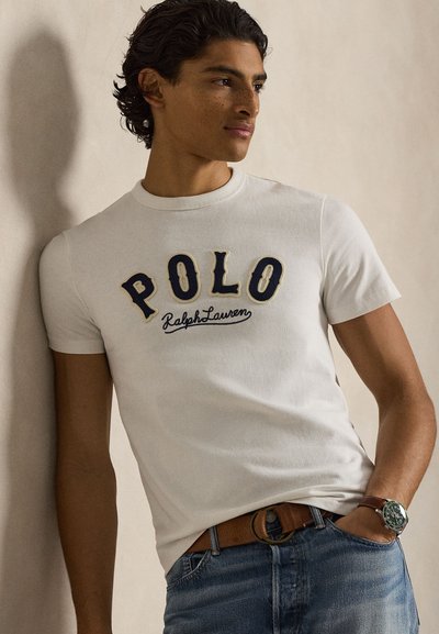 Νεαρός με σκούρα σγουρά μαλλιά που φοράει λευκό μπλουζάκι "POLO Ralph Lauren", καφέ ζώνη, μπλε τζιν και πράσινο ρολόι χειρός, στηριζόμενος σε έναν τοίχο.