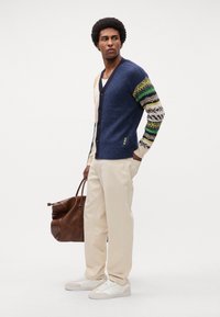 Strikcardigan med blå krop og mønstrede ærmer i grønne, hvide og brune nuancer. Båret med beige bukser og hvide sneakers, mens der holds en brun taske.