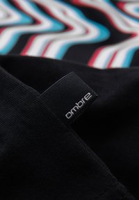 Tissu noir avec une étiquette noire cousue portant la mention « ombre » sur un fond à motif zigzag rouge, blanc et bleu flou.