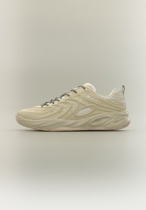 Beiger Sportschuh mit wellenförmigem, strukturiertem Design, grauen Schnürsenkeln und dicker gepolsterter Sohle auf einfarbig hellem Hintergrund.