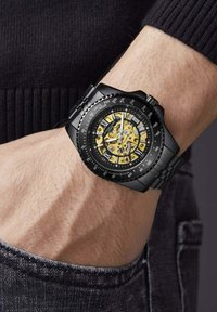 Zwarte roestvrijstalen horloge met een skeleton dial die gele accenten toont, met tachymeter-markeringen en een band met een gepolijste afwerking.