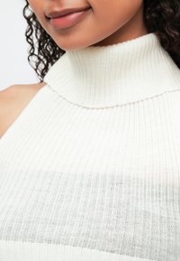 Nærbilde av en person som har på seg en hvit ribbete utenermet turtleneck genser, som viser leppene, haken og en del av krøllete hår.