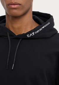 Svart hettegenser laget av myk bomull, med snor i hetta, ribbestrikkede mansjetter og EA7 Emporio Armani-logo brodert på kragen.