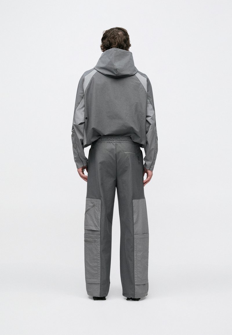 Grå udendørsjakke med hætte og matchende bukser. Begge har strukturerede paneler og cargolommer. Glat stof med en afslappet pasform.