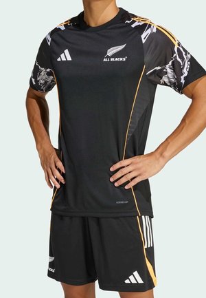 Osoba ubrana w czarną koszulkę rugby All Blacks i pasujące szorty z białymi i pomarańczowymi akcentami, stoi z rękami na biodrach.