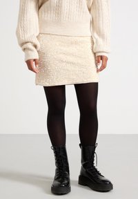 Pärlad krämfärgad mini kjol med en figurnära design, ihop med svarta strumpbyxor och svarta snörda kängor. Texturerad stickad tröja ovanpå.