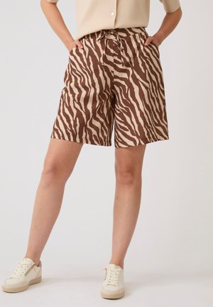 Persoon die bruine en beige zebraprint shorts draagt met handen in de zakken en witte sneakers, staand tegen een effen achtergrond.