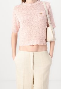Strickpullover mit kurzen Ärmeln in sanftem Pink, ausgestattet mit einem gerippten Ausschnitt und einem kleinen dekorativen Logo auf der Vorderseite, kombiniert mit hellbeigen Hosen.